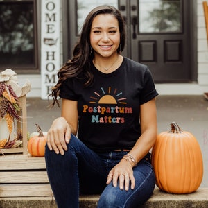 May include: A woman wearing a black t-shirt with the text "Postpartum Matters" and a colorful sunset graphic. She is sitting on a wooden bench with pumpkins on either side of her.