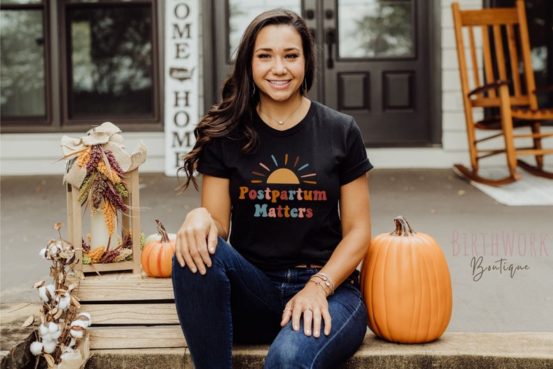 May include: A woman wearing a black t-shirt with the text "Postpartum Matters" and a colorful sunset graphic. She is sitting on a wooden bench with pumpkins on either side of her.