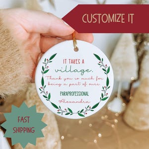 May include: A white ceramic ornament with a gold string for hanging. The ornament has a green and red floral border and text that reads "IT TAKES A village. Thank you so much for being a part of ours. PARAPROFESSIONAL Alexandra."