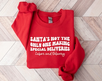 May include: A red sweatshirt with white text that reads "SANTA'S NOT THE ONLY ONE MAKING SPECIAL DELIVERIES" and "Labor and Delivery". The sweatshirt is folded on a light wood-grain surface.
