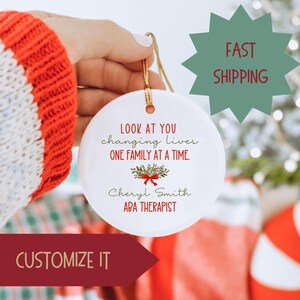 May include: White ceramic ornament with the text "LOOK AT YOU changing lives ONE FAMILY AT A TIME." and "Cheryl Smith ABA THERAPIST". The ornament is decorated with a red bow and green leaves.