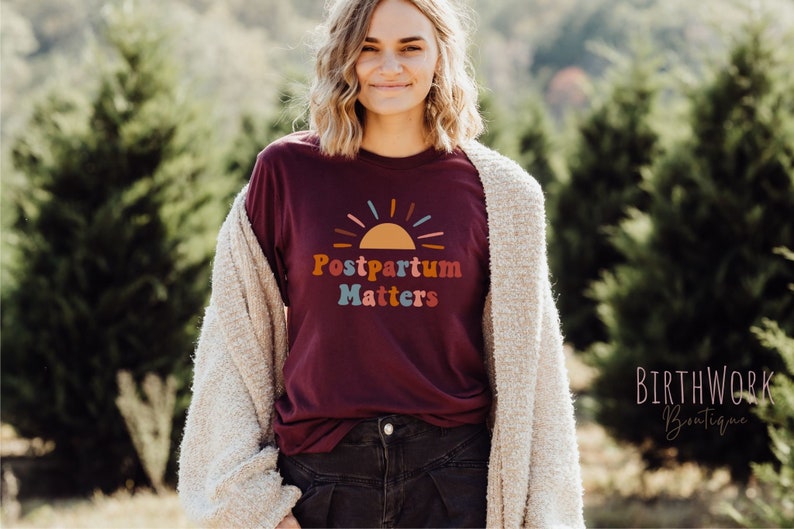 May include: A maroon t-shirt with a colorful graphic of a sunrise and the text "Postpartum Matters".