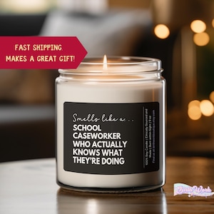 May include: A white candle with a black label that reads "Smells like a... School Caseworker Who Actually Knows What They're Doing". The candle is in a clear glass jar and is lit.