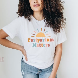 May include: A white t-shirt with a colorful graphic of a sun and the text "Postpartum Matters".