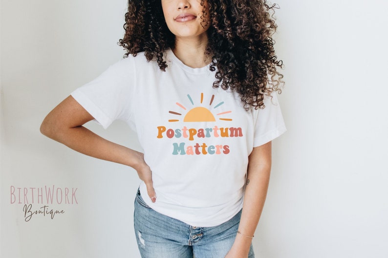May include: A white t-shirt with a colorful graphic of a sun and the text "Postpartum Matters".