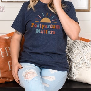 May include: A navy blue t-shirt with a colorful graphic of a sun and the text "Postpartum Matters".