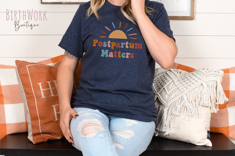 May include: A navy blue t-shirt with a colorful graphic of a sun and the text "Postpartum Matters".