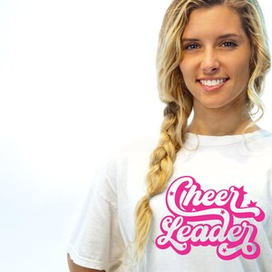 May include: White t-shirt with the words "Cheer Leader" in a pink, stylized font. The text is accented with stars. The shirt is worn by a person with blonde hair in a braid.