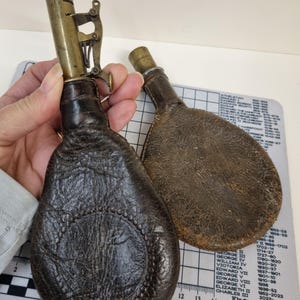 Op de afbeelding: Twee antieke leren poederflessen. Eén fles wordt door een hand vastgehouden en toont een donkerbruine, getextureerde lederen behuizing met een messing tuit en mechanisme. De andere fles is lichtbruin. De achtergrond bevat een liniaal en een lijst met Engelse monarchen.