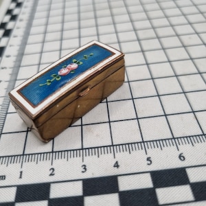 May include: A small, rectangular brass box with a hinged lid. The lid has a blue enamel panel with a white border, decorated with a floral design in pink and green. The box is on a grid-patterned surface.
