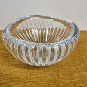 Könnte beinhalten: Eine klare Glasschale mit vertikalen Rippendetails. Die Schale hat eine abgerundete Form und steht auf einer Holzoberfläche. Das Glas ist transparent, so dass Licht hindurchscheinen kann, und das gerippte Design sorgt für visuelles Interesse.