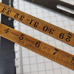 Könnte beinhalten: Nahaufnahme von Vintage-Holzlinealen und einem Metallwerkzeug auf einer karierten Oberfläche. Ein Lineal ist in Zentimetern markiert und zeigt Messungen von 10 bis 20 cm. Das andere Lineal zeigt Messungen von 74 bis 84 cm.