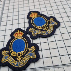 Könnte beinhalten: Zwei blaue und goldene gestickte Patches mit dem Text "Ralph Lauren" auf einem marineblauen Hintergrund. Die Patches zeigen eine Krone und einen blauen Kreis mit einem goldenen Design.