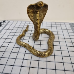 May include: A brass cobra statue with its hood raised, displaying intricate scale detail. The snake's body is coiled, and the statue has a golden colour. It is positioned on a white and black grid, creating a contrast.