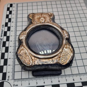 May include: A silver-toned teddy bear picture frame with a velvet backing. The frame has a round opening for a photo and a textured bear face.