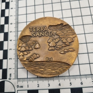 以下が含まれることがあります： 上部に「TERRA SANCTA」の文字が入った、丸いブロンズ色のメダル。 メダルには、山、水、建物のある風景が描かれています。 メダルの直径は約7.6cmです。