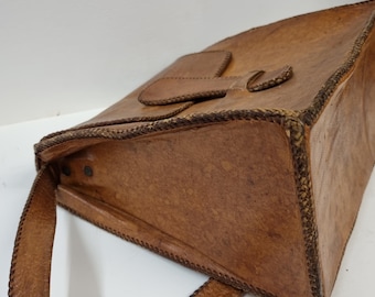 May include: A brown leather tote bag with a rectangular shape and a handle. The bag features a flap closure with a decorative design and stitched edges. The leather has a textured appearance.