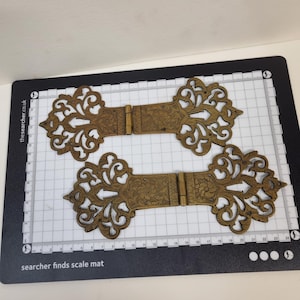 Puede incluir: Dos bisagras de latón antiguo con intrincados grabados florales y diseños ornamentales de volutas caladas en cada extremo. Las bisagras están colocadas sobre una alfombrilla de rejilla blanca y negra con medidas en centímetros, mostrando 24 cm.