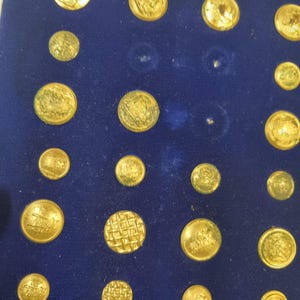 Può includere: Una collezione di bottoni dorati di varie dimensioni, esposti su una superficie di velluto blu scuro. I bottoni presentano disegni in rilievo, tra cui stemmi e motivi. La disposizione suggerisce un oggetto vintage o da collezione.