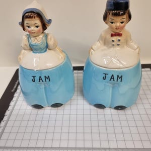 May include: Two ceramic jam jars shaped like figures in blue outfits. The left figure wears a white bonnet and blue dress, while the right figure wears a blue cap and white jacket with a red bowtie. Both jars feature the word JAM in black letters.