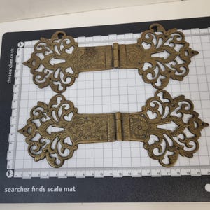May include: Two antique-style brass hinges featuring intricate scrollwork and floral engravings. Each hinge measures approximately 8.5 inches in total length when laid flat on a grid mat with clearly visible centimeter markings.