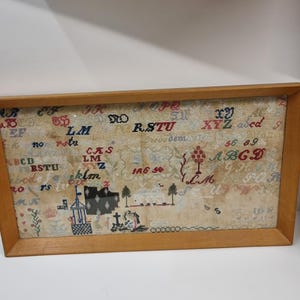 以下が含まれることがあります： 薄いタン色の背景に額装されたアンティークのサンプラー。 サンプラーには、刺繍された文字、数字、井戸、木、建物の小さなイラストが描かれています。 フレームは明るい茶色の木材で作られています。