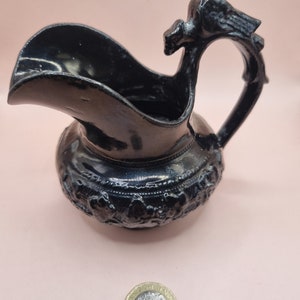 May include: A small, black ceramic pitcher with an ornate design and a bird-shaped handle. The pitcher is about the size of a British pound coin.