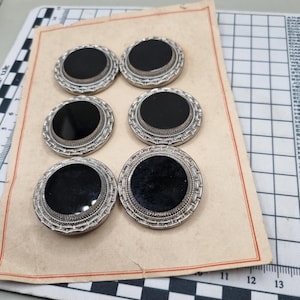 May include: Six vintage buttons with a black centre and a silver-coloured, ornate border. The buttons are arranged on a piece of aged paper, suggesting a classic or antique style. The buttons are round and have a detailed, textured design.