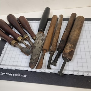 Op de afbeelding: Een verzameling van zeven diverse handgereedschappen met houten handvatten, waaronder beitels, een paletmes en metalen snijgereedschap, gerangschikt op een witte rastermat met een zichtbare schaalverdeling tot 20 centimeter.