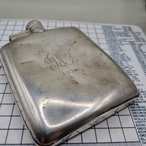 Peut inclure: Une flasque rectangulaire en argent avec un bouchon à vis. La flasque est gravée des initiales "GS" et de l'année "1916". La flasque a un aspect légèrement usé et mesure environ 13 cm de long.