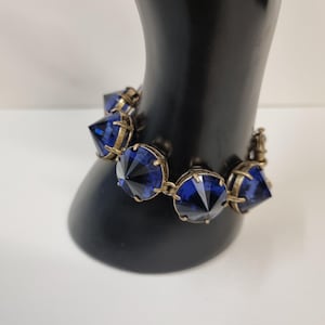 Op de afbeelding: Een armband met facetgeslepen, diepblauwe edelstenen, gezet in een goudkleurig metaal. De armband wordt getoond op een zwarte mannequinhand, waarbij het ontwerp en de kleur van de sieraden worden benadrukt.