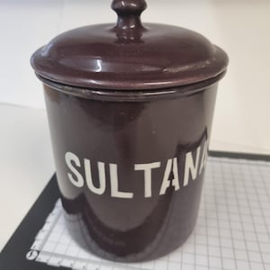 以下が含まれることがあります： 蓋と小さなノブが付いた、濃い茶色のセラミックジャー。「SULTAN」の文字が白で側面に印刷されています。ジャーは円筒形で、キッチン用の収納容器のようです。