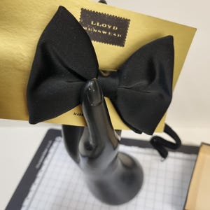 Pode incluir: Uma gravata borboleta de cetim preto liso presa a um cartão de exibição dourado. O cartão apresenta uma etiqueta retangular escura com o texto LLOYD MENSWEAR impresso em dourado. A gravata borboleta tem um acabamento liso e um nó central.