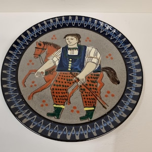 May include: A decorative ceramic plate featuring a folk art illustration. The central image depicts a person in traditional attire riding a horse. The plate has a blue border with a repeating pattern.