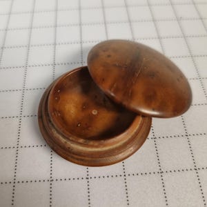 May include: A small, round wooden box with a lid. The box and lid are a warm brown color, with visible wood grain. The lid is slightly ajar, revealing the interior of the box. The box appears to be handmade.