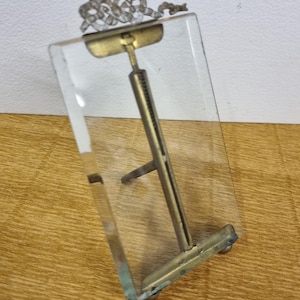 Puede incluir: Un marco de fotos de vidrio transparente con una parte superior decorativa de metal y un soporte de color latón. El marco es rectangular e inclinado, con un soporte de metal en la parte posterior. La parte superior presenta un diseño ornamental en forma de cinta.