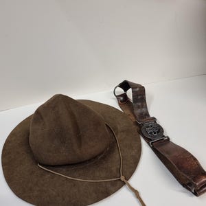 Könnte beinhalten: Ein brauner Filzhut mit breiter Krempe und Kinnriemen, neben einem braunen Ledergürtel mit Metallschnalle. Der Hut hat eine mittelbraune Farbe, und der Gürtel weist Gebrauchsspuren auf.
