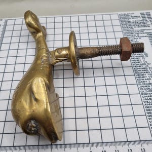 Puede incluir: Un aldaba de puerta de latón con forma de delfín con un perno roscado y una tuerca. El delfín tiene aletas detalladas y una superficie texturizada. El perno se extiende desde una placa circular y la tuerca está oxidada.