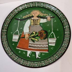 May include: A round, hand-painted decorative plate with a green background and a detailed border. The plate features a folk art illustration of a woman carrying buckets and holding a flower. The woman is wearing a traditional dress with orange, brown, and white accents.
