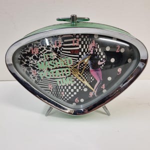 May include: A green and chrome alarm clock with a black face and a white and black chequered pattern. The clock face also features a woman in a pink dress and the text "It's mashed potato time".