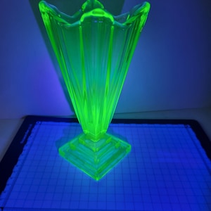 May include: A striking, neon green glass vase with a flared top and a geometric base. The vase has a distinctive shape and is illuminated, creating a bright glow. The vase is likely vintage or antique.