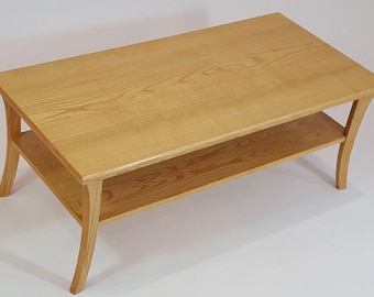 May include: A rectangular light brown wooden coffee table featuring a smooth top surface and a lower shelf for storage. The table is supported by four curved legs that flare outward, showcasing a natural wood grain pattern throughout the structure.