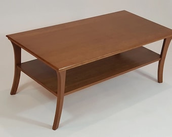 May include: A rectangular coffee table crafted from medium-toned brown wood. The table features a smooth, flat top surface and a lower shelf for storage, supported by four curved legs that extend outwards from the corners of the frame.