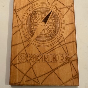 Pode incluir: Uma placa de madeira com um design de b&uacute;ssola e as palavras "ONE PIECE" em uma fonte estilizada. A b&uacute;ssola &eacute; cercada por um padr&atilde;o de teia de aranha.
