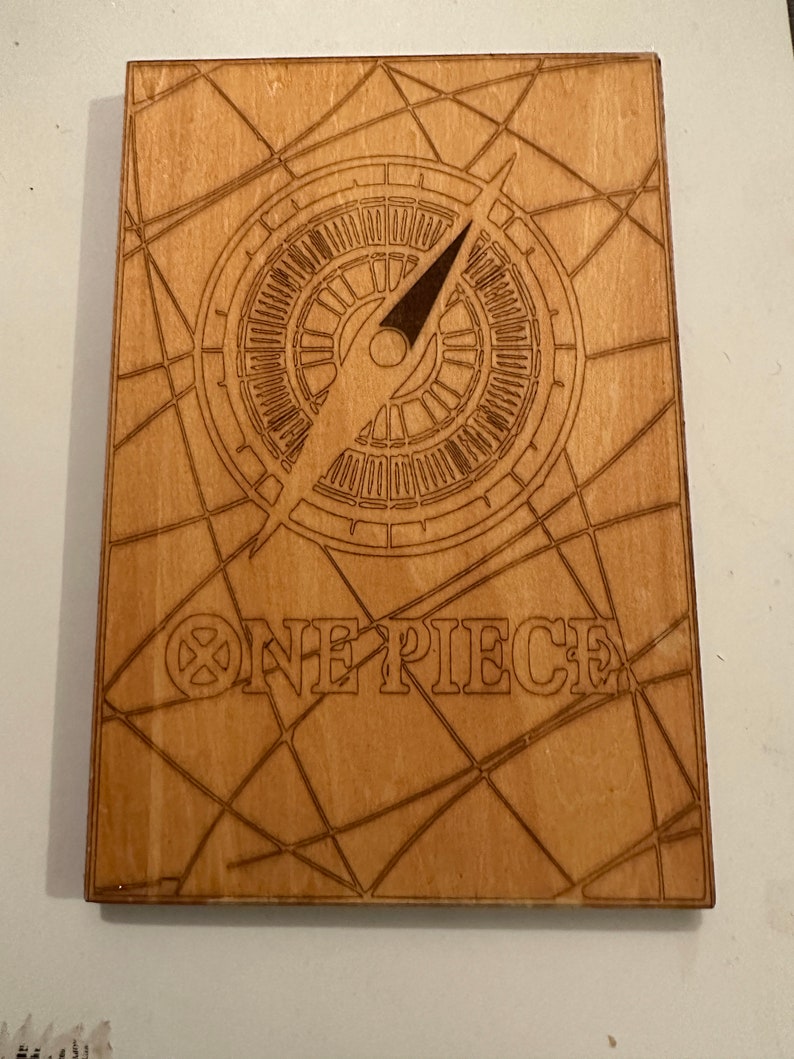 Pode incluir: Uma placa de madeira com um design de b&uacute;ssola e as palavras "ONE PIECE" em uma fonte estilizada. A b&uacute;ssola &eacute; cercada por um padr&atilde;o de teia de aranha.