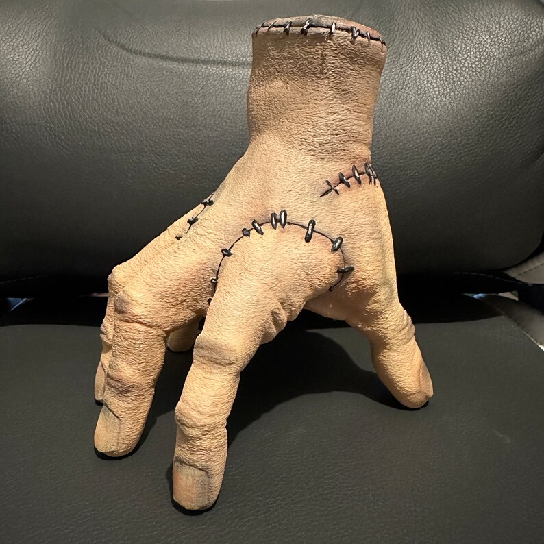 May include: A beige, stitched, sculpted hand with four fingers. The hand is made of a textured material and has black stitching around the wrist and fingers.
