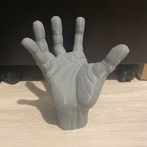 May include: A 3D printed gray hand sculpture with all five fingers extended. The hand is posed as if it is reaching up.