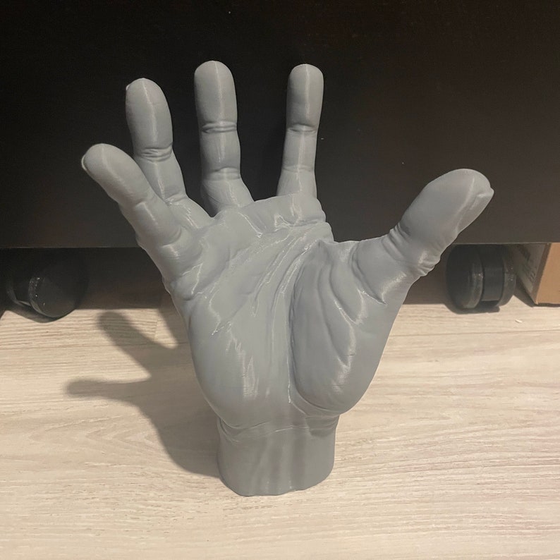 May include: A 3D printed gray hand sculpture with all five fingers extended. The hand is posed as if it is reaching up.
