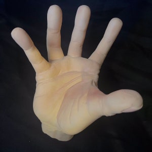 May include: A close-up of a pale, flesh-toned, realistic-looking hand with five fingers extended, palm facing the camera. The hand is made of a smooth, slightly textured material.