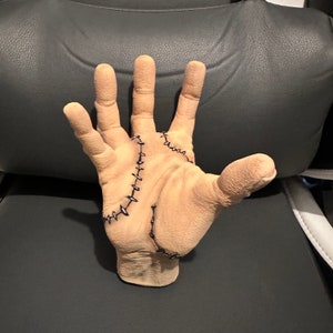 May include: A realistic-looking, flesh-toned hand with stitched seams across the palm and fingers. The hand is posed with fingers spread apart, as if reaching out.